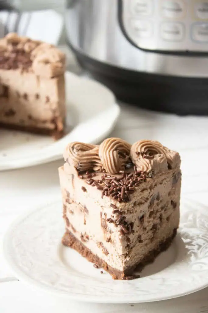 Close up of a slice of Instant Pot Chocolate Chip Cheesecake on a white plate, remainder of cheesecake in the background together with the Instant Pot appliance.