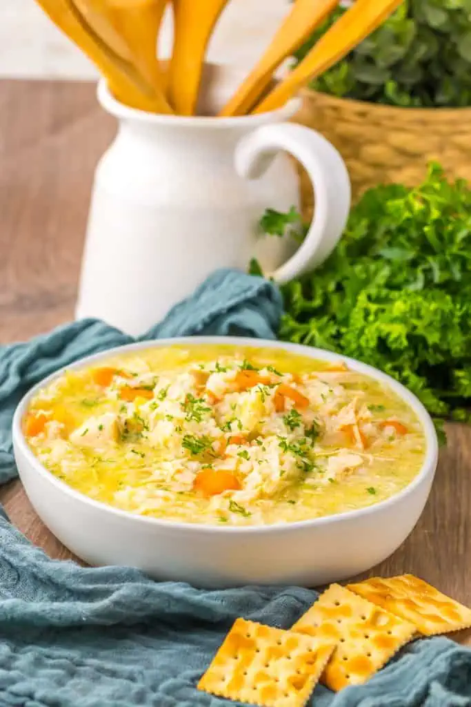 Slow cooker chicken and rice soup in a white serving bowl with a blue kitchen cloth, fresh parsley, crackers, and cooking utensils in the background.