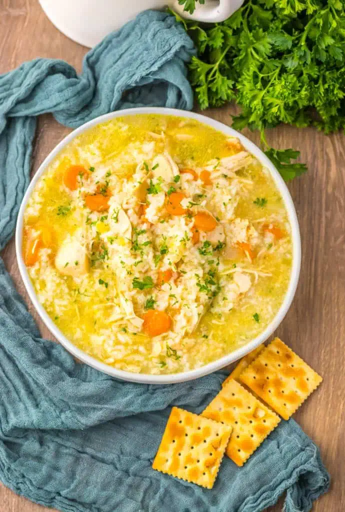 Overhead view of Slow cooker chicken and rice soup in a white serving bowl with a blue kitchen cloth, fresh parsley, crackers, and cooking utensils.