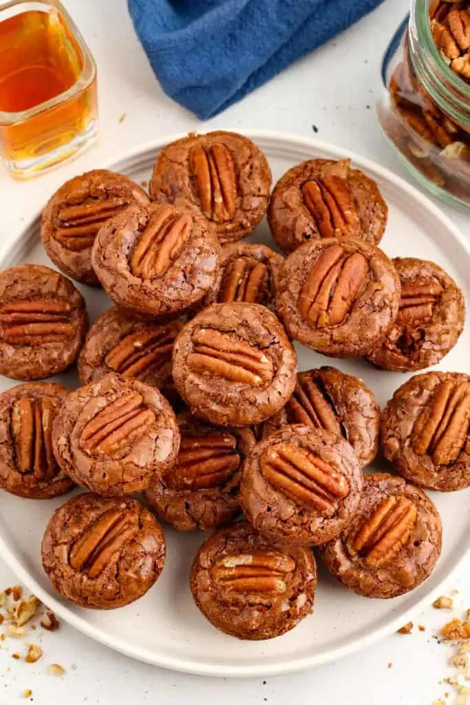 Bourbon pecan brownie bites piled on a white serving plate, jar of pecans, shot glass of bourbon, and blue cloth.