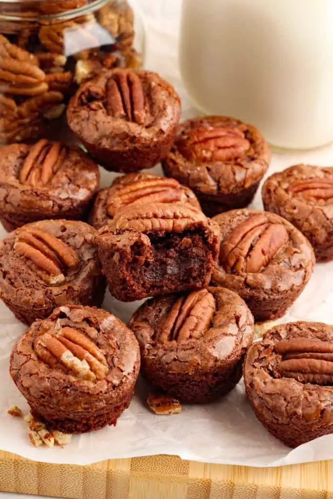 Close up of bourbon pecan brownie bites piled on a parchment paper-lined wooden board, jar of pecans, glass of milk.
