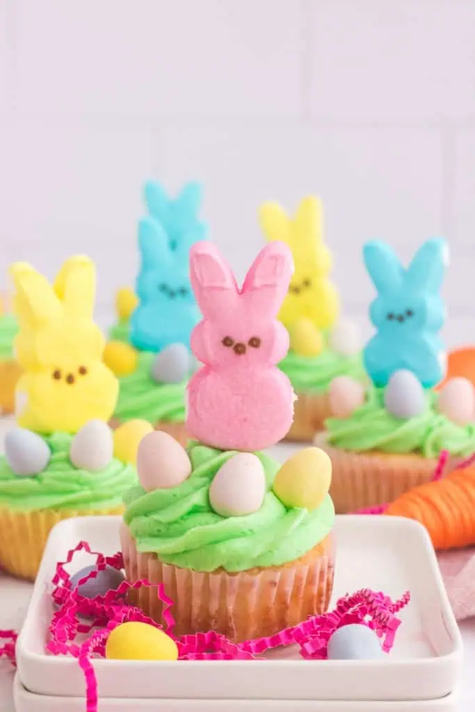 Close up of a Peeps cupcake in a small square dish with decorative shredded paper, more peeps cupcakes in the background.