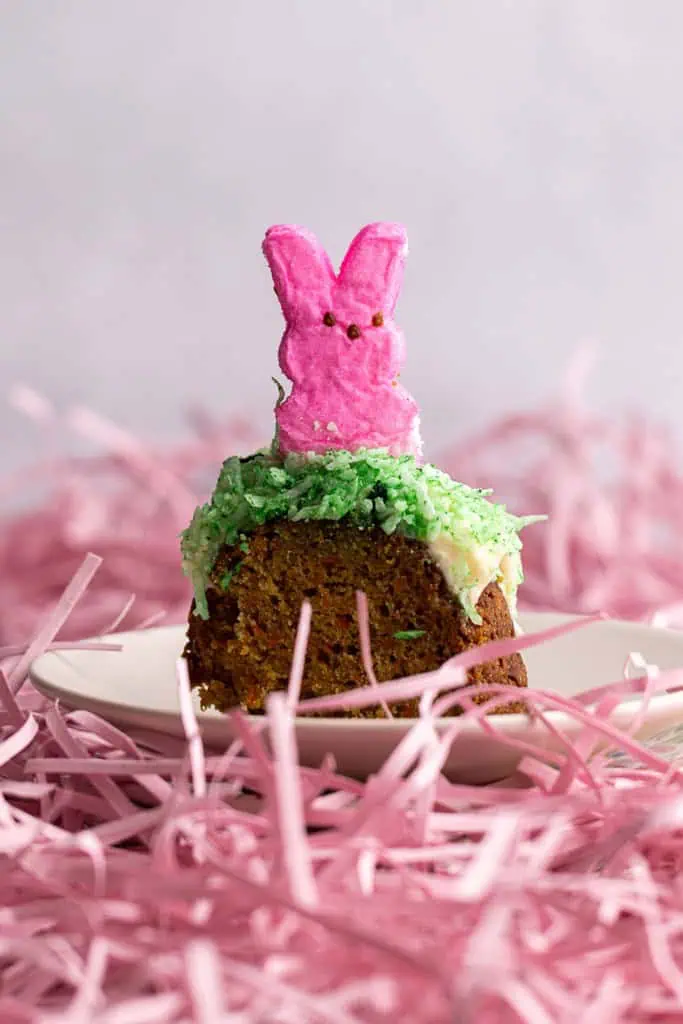 Close up of a slice of Peeps carrot bundt cake on a white side plate with pink decorate shredded paper scattered around.