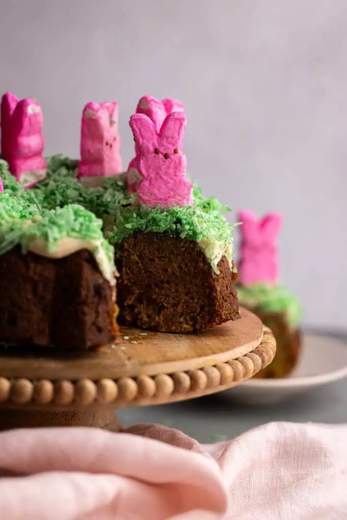Peeps carrot bundt cake on a cake stand with cream cheese frosting, green-tinted coconut grass, and pink marshmallow peeps.