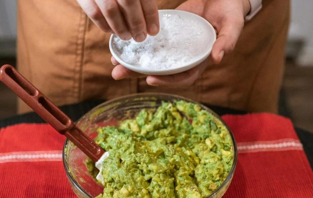 Hands preparing fresh guacamole with avocado and salt in kitchen.