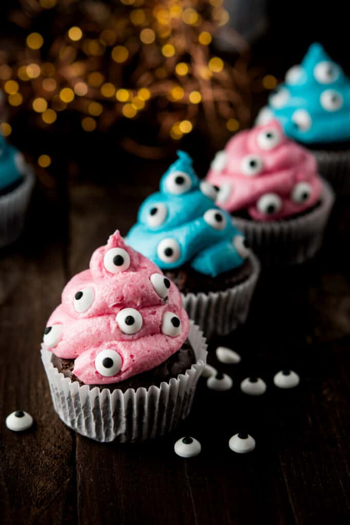a row of blue and pink iced monster cupcakes with candied eyes on table