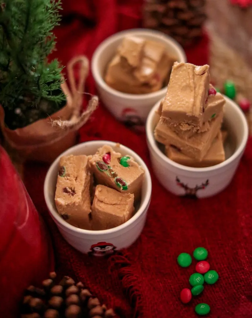 christmas peanut butter fudge in white christmas containers on a red background with red and green m&ms scattered