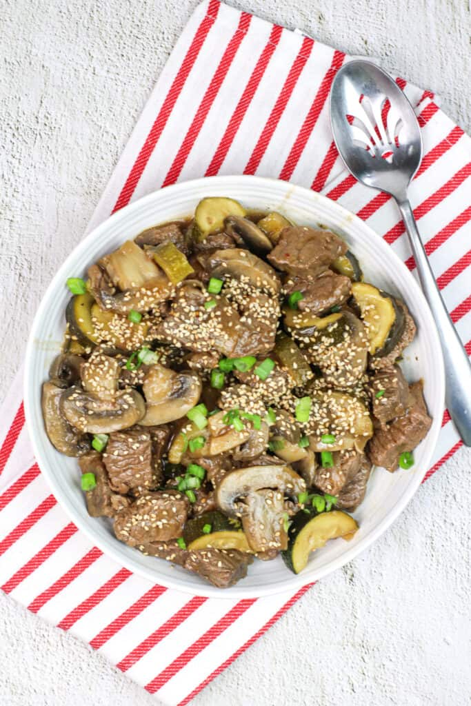 instant pot hibachi steak bites in a white bowl and a red and white linne with a slotted spoon garnished with sesame seeds