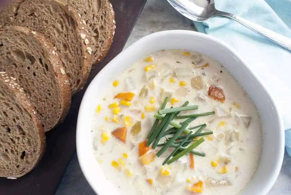 horizontal image of corn chowder in a white bowl with sliced wheat bread on a brown board