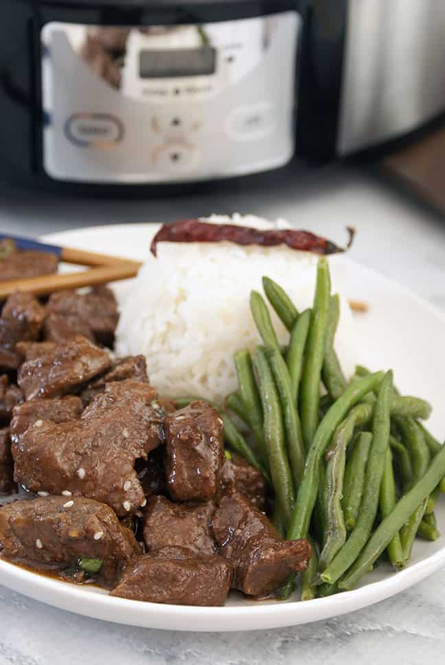 side view of slow cooker korean beef on a white plate next to a crockpot