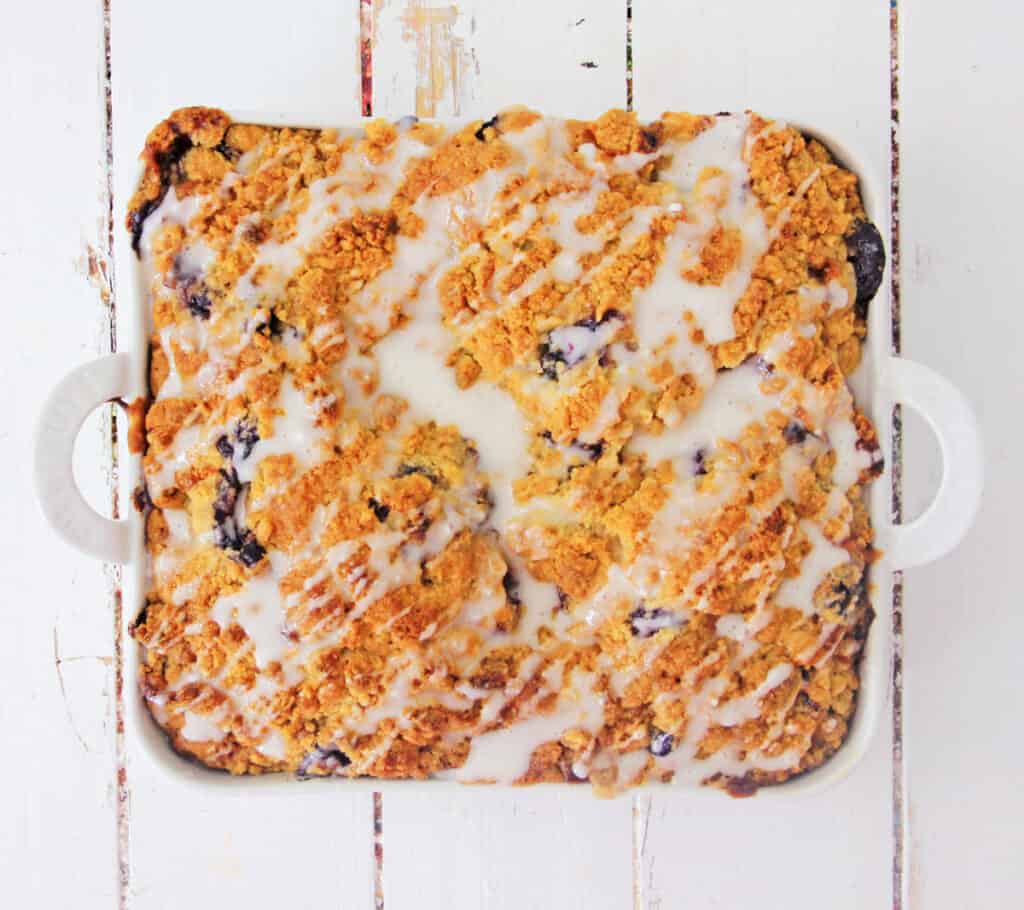 overhead shot of a blueberry crumb coffee cake with a glaze drizzle in a white ceramic baking dish