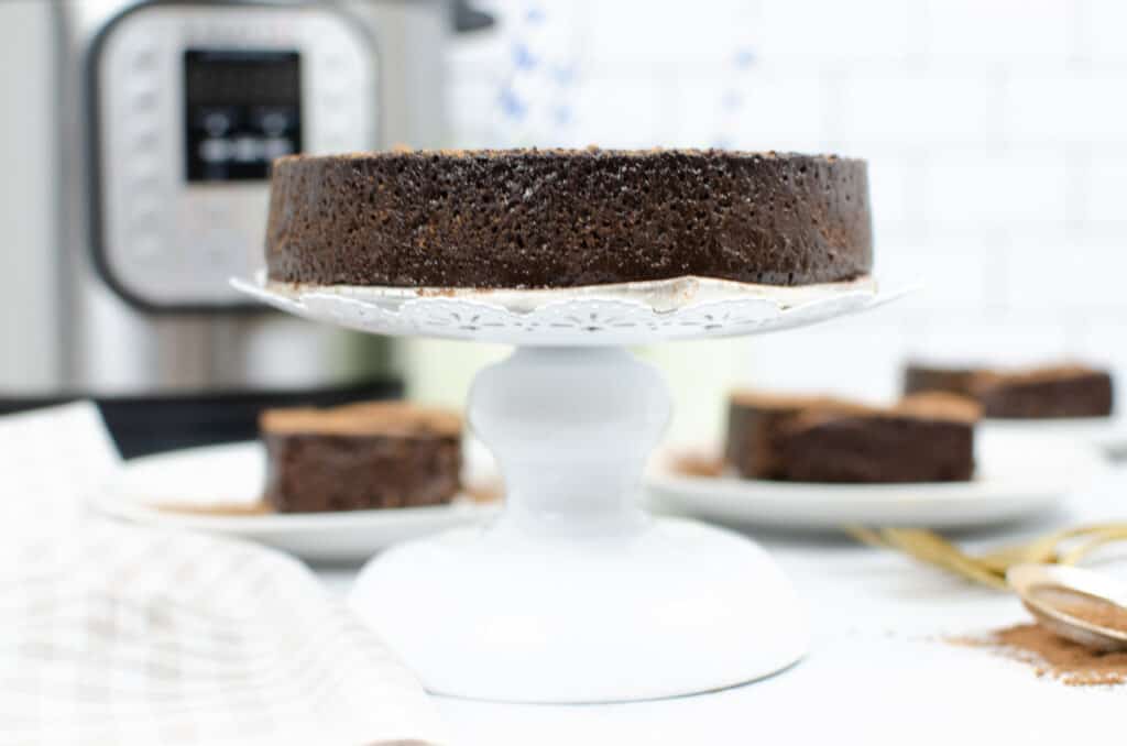 instant pot flourless chocolate cake in front of a pressure cooker on a white ceramic cake platter