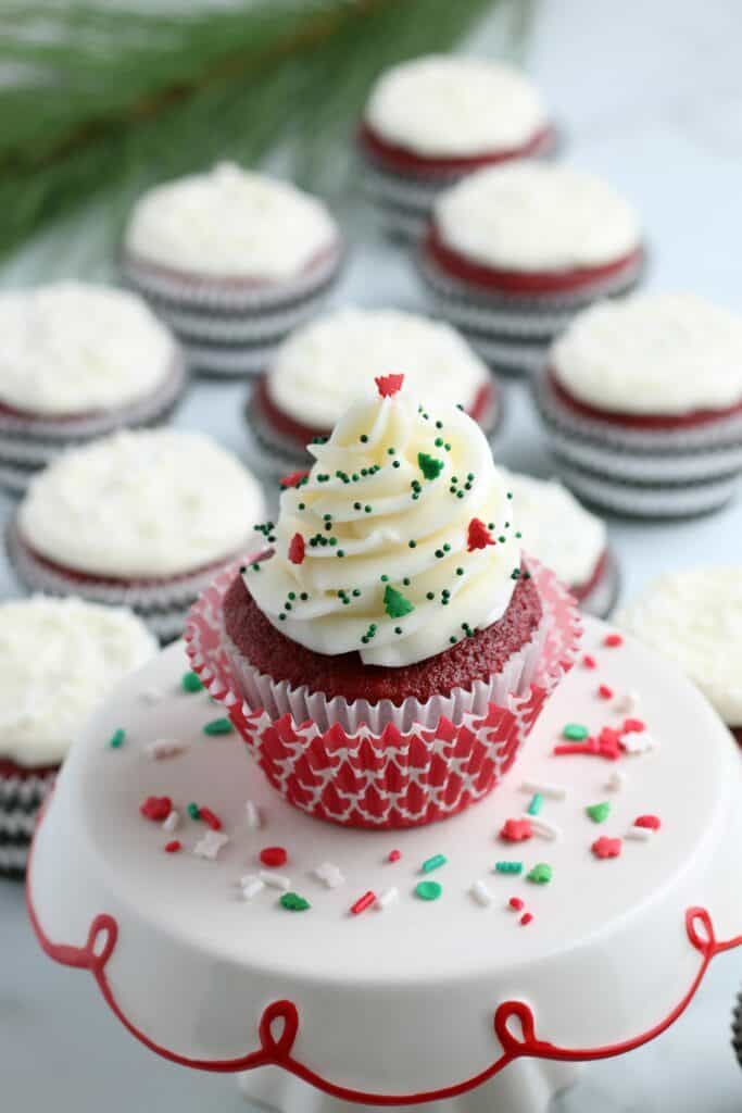 a christmas red velvet cupcake recipe with christmas sprinkles on a white and red ceramic cake stand