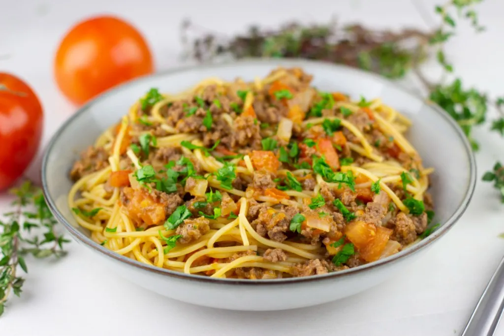 a yummy serving of a one pot taco spaghetti on a brown rimmed stone plate with fresh tomatoes and herbs in the background