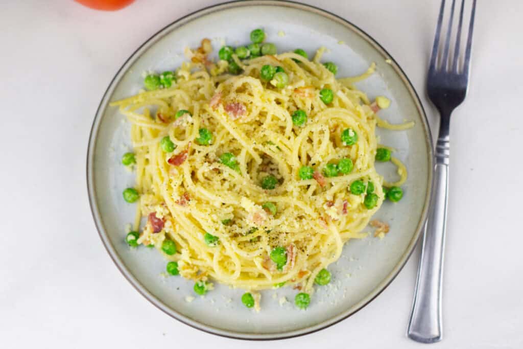 flay lay view of a mound of pasta carbonara with fresh peas sprinkled around next to a fork on a white table background