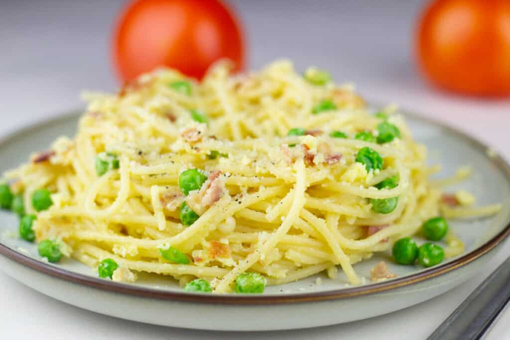 an easy to make pasta carbonara recipe that's served on a white stone speckled plate with a brown rim and tomatoes in the background