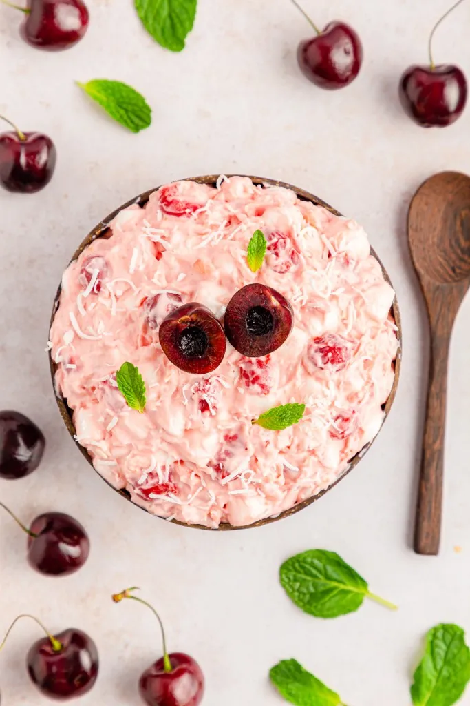 Overhead hero shot of a mound of cherry fluff salad served in a dark wooden bowl with fresh cherries and wooden spoon