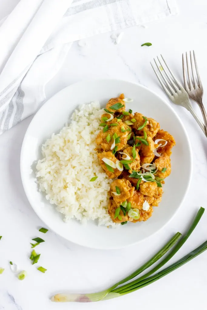 overhead shot of homemade copycat panda express orange chicken with cooked rice and diced green onions on a white plate with 2 metal forks.