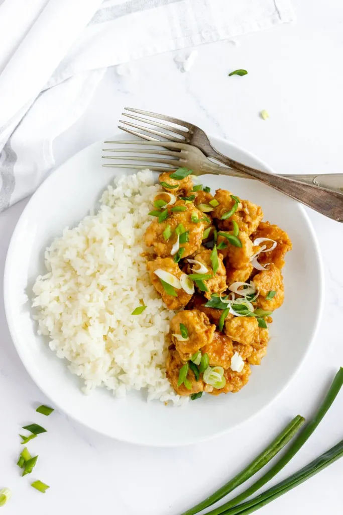 overhead shot of homemade copycat panda express orange chicken with cooked rice and diced green onions on a white plate.