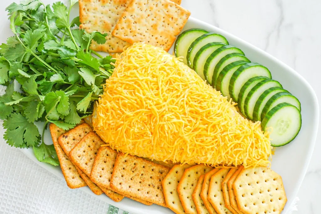 overview of easter cheese ball with crackers and cucumbers on a rectangular shaped white plate