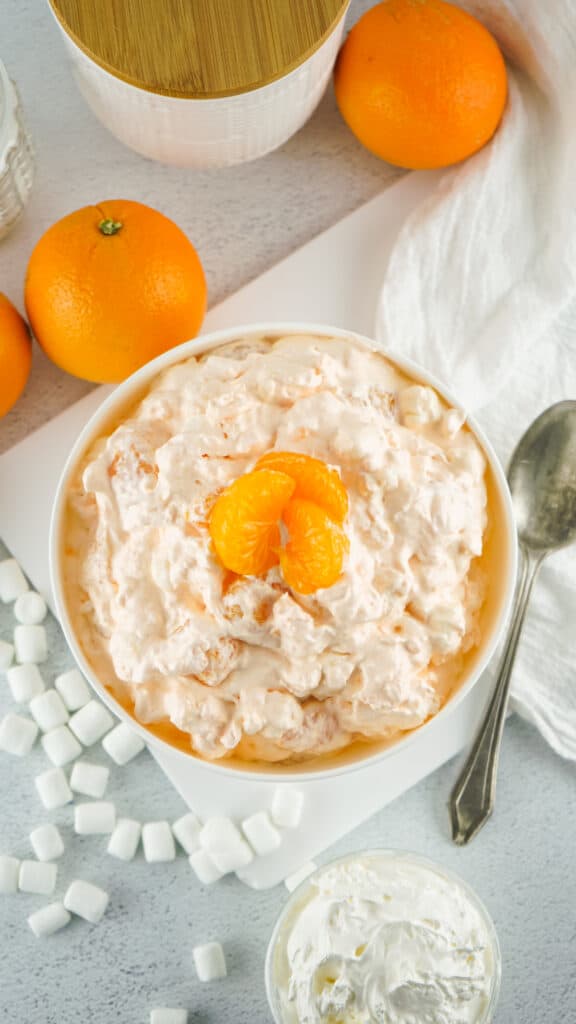 orange fluff salad in a white bowl surrounded my oranges and mini marshmallows