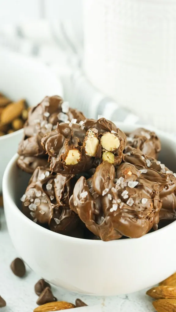 a white ceramic bowl filled with sea salt chocolate almond clusters with almonds and chocolate chips scattered around and an open cluster showing almonds