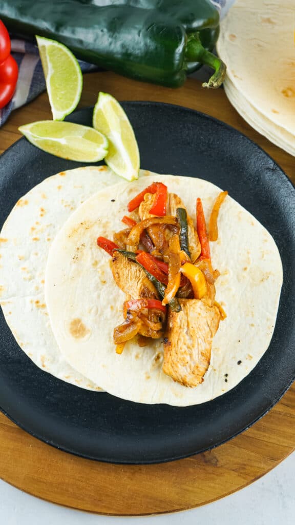 overhead image of an open face chicken fajitas recipe on tortillas, a black plate, and placed on a wooden round board.