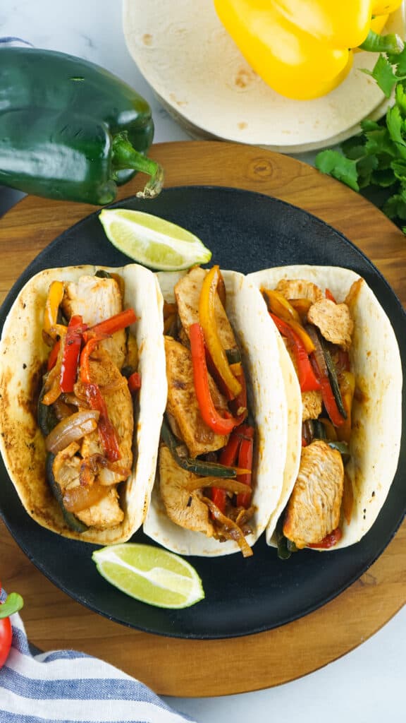 overhead photo of 3 skillet chicken fajitas on a black plate and round board with green poblano peppers and sliced limes int he background