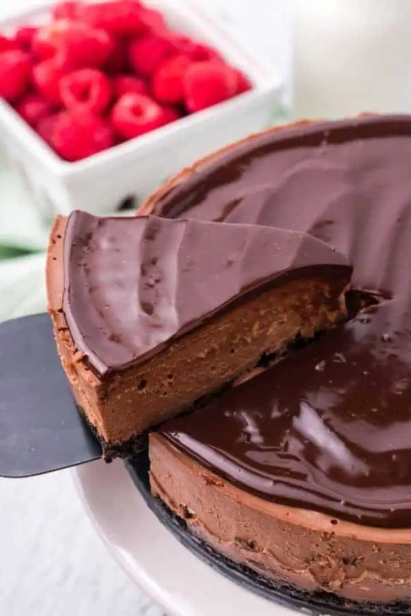 a slice of chocolate cheesecake being lifted from whole cheesecake with raspberries in background