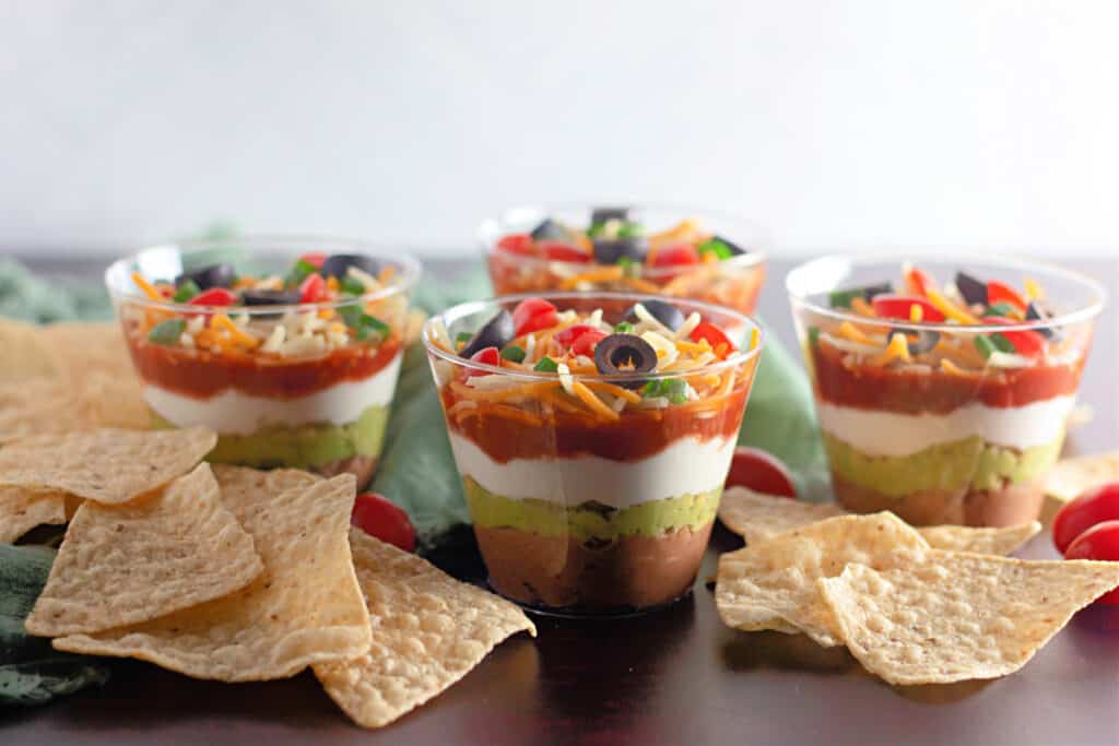 Assembled 7 Layer Dip Cups with tortilla chips on a dark countertop.