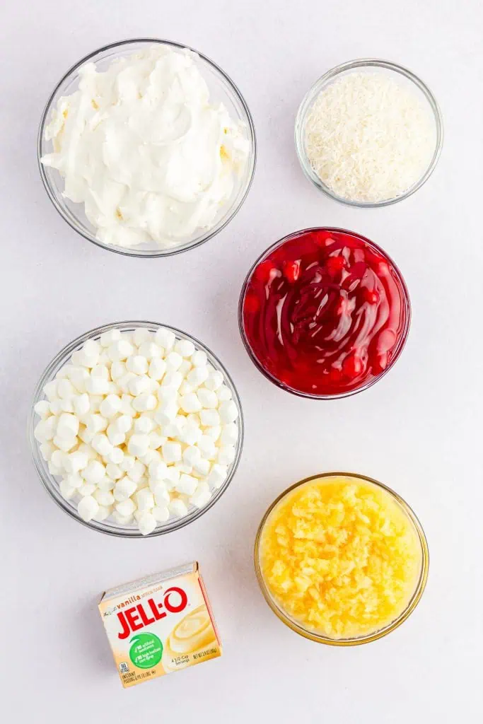 ingredients need for vintage cherry fluff salad with bowls of mini marshmallows, crushed pineapple, cherry pie filling, shredded coconut and box of pudding