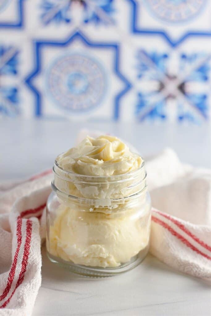 Glass jar of homemade butter with a white a red striped kitchen cloth on a white marble countertop, blue and white decorative tiles in the background.
