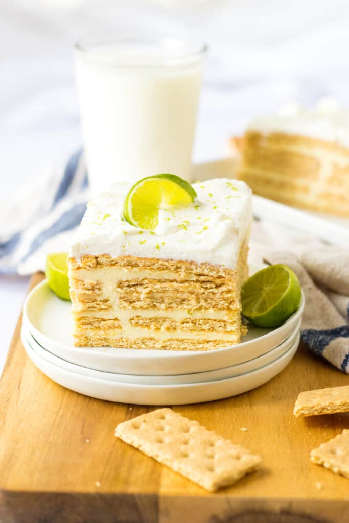 Slice of key lime icebox cake on stacked white plates, glass of milk and cream kitchen cloth in the background, on a wooden kitchen board.