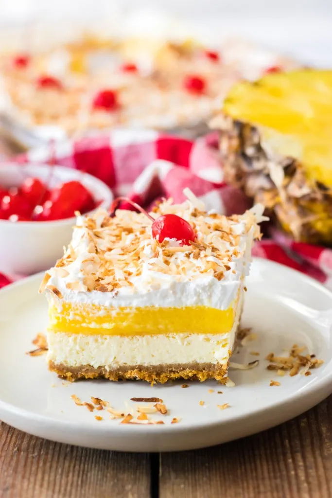 A slice of Pina Colada Cheesecake Bars dessert on a white serving plate, bowl of maraschino cherries, baking tray with Pina Colada Cheesecake in the background.