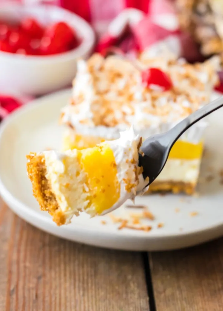 Metal fork holding a piece of Pina Colada Cheesecake bars dessert, slice of cheesecake on a white plate, small bowl of maraschino cherries, piece of cut pineapple, and red and white checkered kitchen cloth in the background.