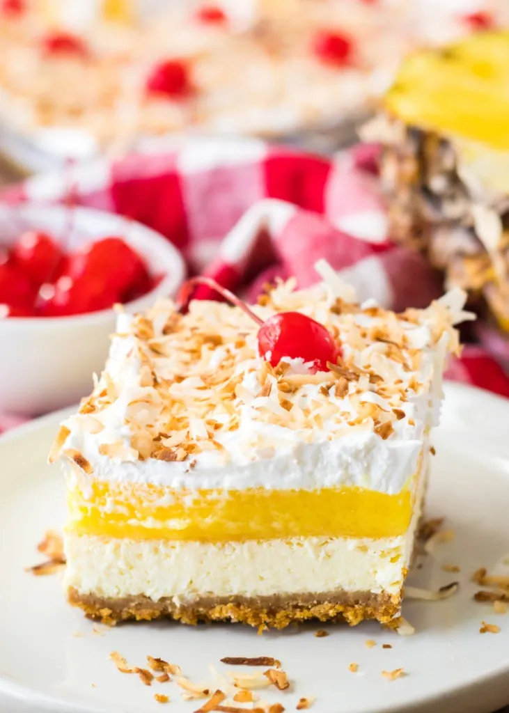A slice of Pina Colada Cheesecake Bars dessert on a white serving plate, bowl of maraschino cherries, baking tray with Pina Colada Cheesecake in the background.