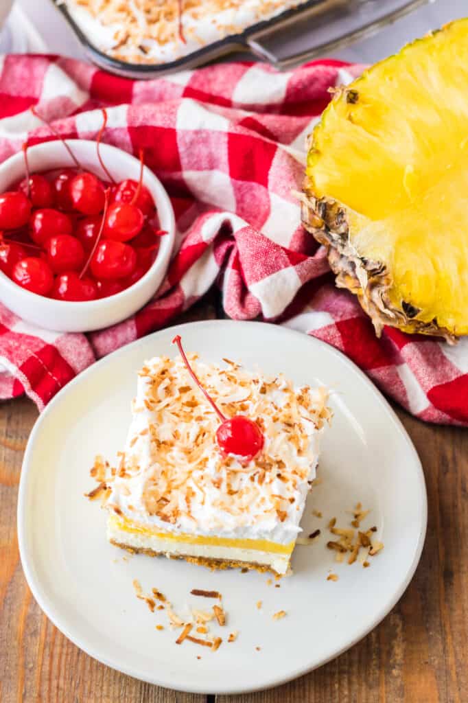 Overhead view of a slice of Pina Colada Cheesecake Bars dessert on a white plate, small bowl of maraschino cherries, freshly cut pineapple, baking tray with remaining Pina Colada cheesecake, red and white checkered cloth.