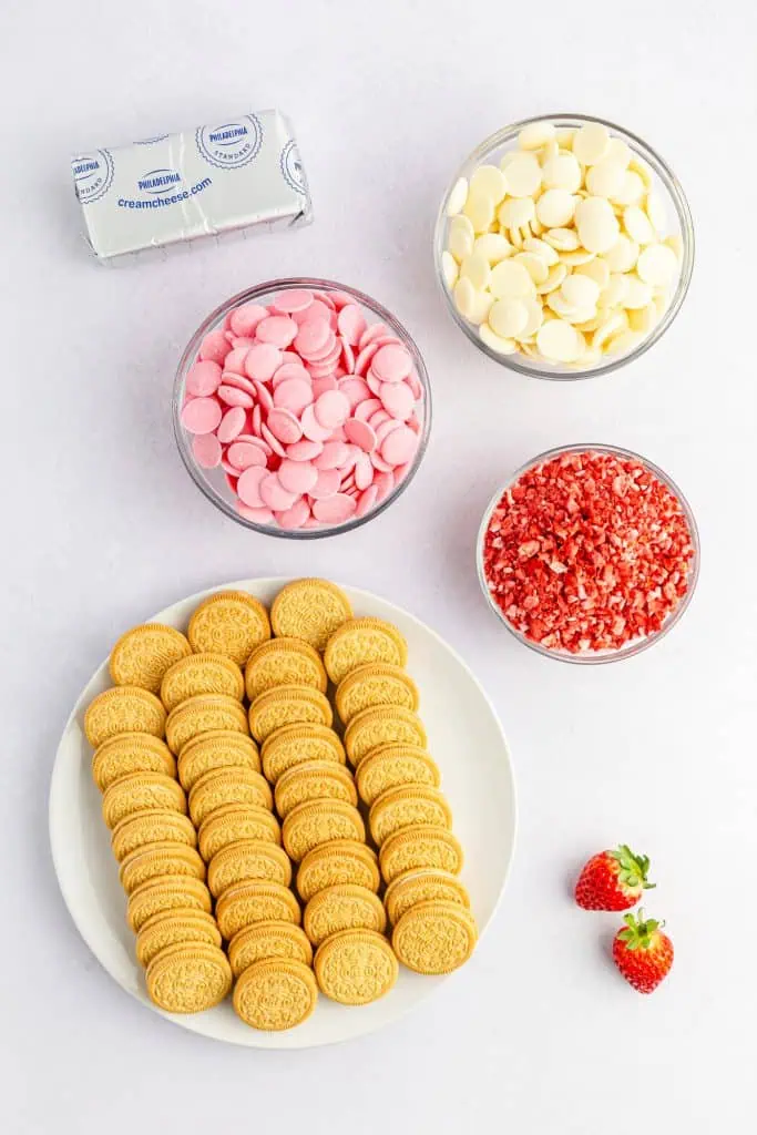 golden oreo cookies, pink melting chocolates, white melting chocolates, strawberry runch and cream cheese ingredients for strawberry shortcake crunch balls on a white background