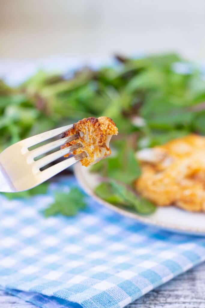 Metal fork holding a piece of air fryer cauliflower steak, serving plate with air fryer cauliflower steak and fresh greens in the background, white and blue checkered table cloth.