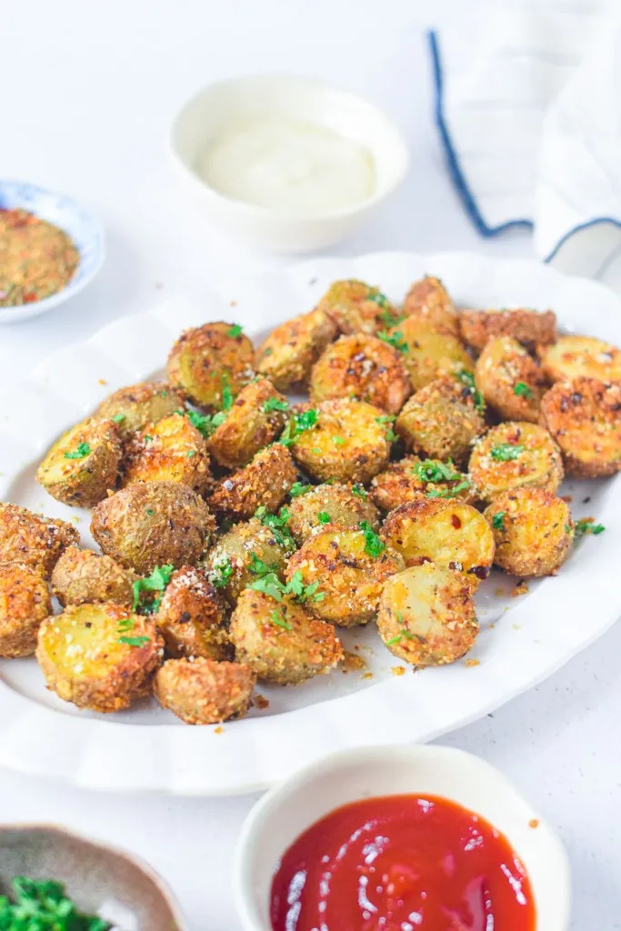 White serving plate with air fryer parmesan potatoes, bowl of ketchup, bowl of seasoning, bowl of fresh herbs, freezer bag, on a white countertop.