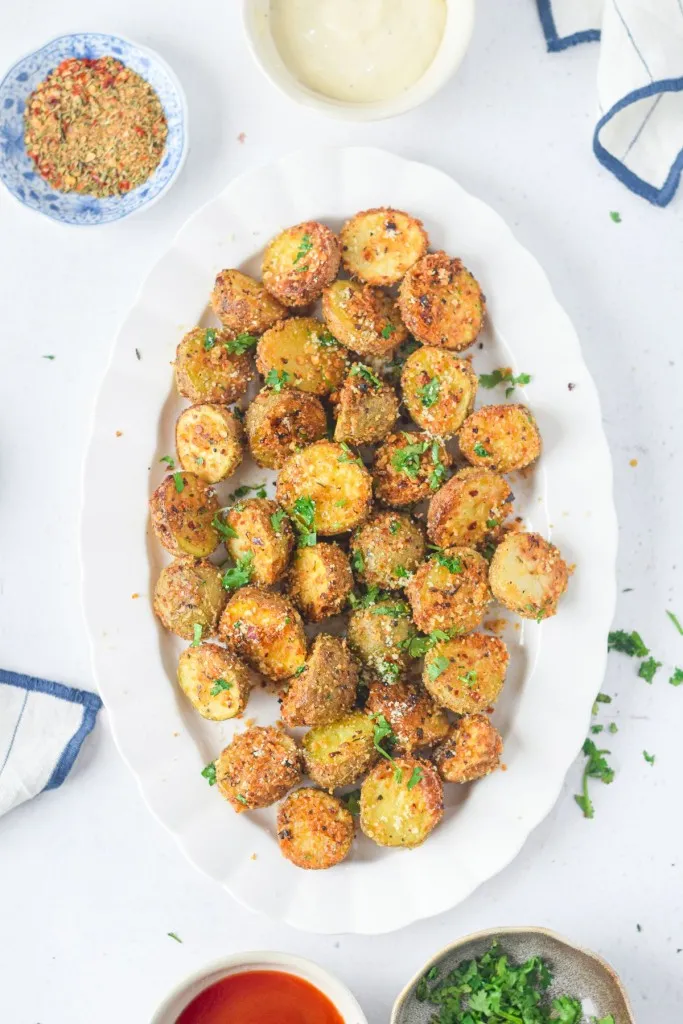 White serving plate with air fryer parmesan potatoes, bowl of ketchup, bowl of seasoning, bowl of fresh herbs, kitchen cloth, on a white countertop.