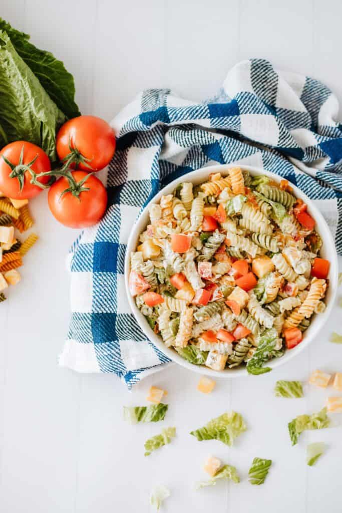 Overhead view of a bowl of BLT Pasta Salad, blue and white checkered kitchen cloth, fresh tomatoes, on a white kitchen countertop.
