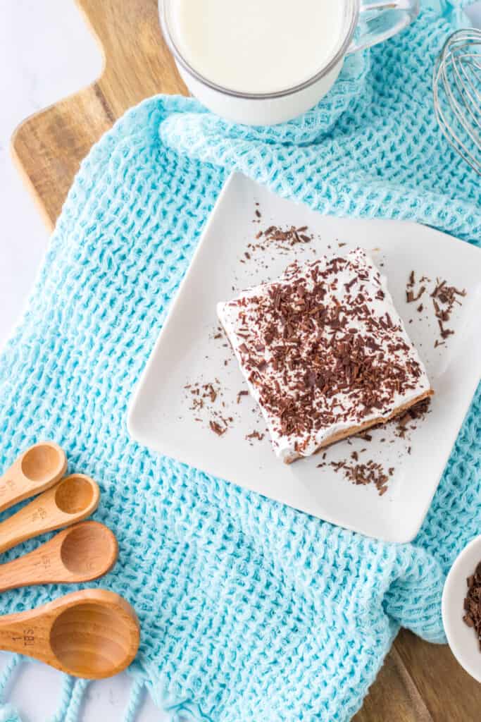 Overhead view of a slice of French Silk Brownies on a white plate, wooden measuring spoons, glass of cold milk, pale blue kitchen cloth, on a wooden kitchen board.