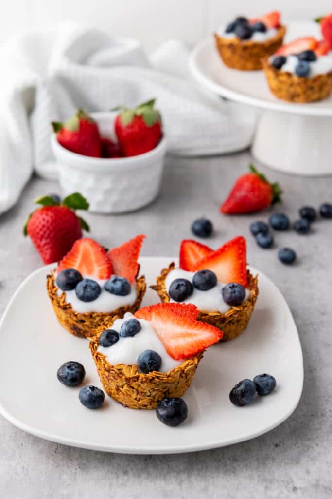 Three granola sups with yogurt on a white serving plate, white ramekin with fresh fruit, cake stand with extra granola cups.