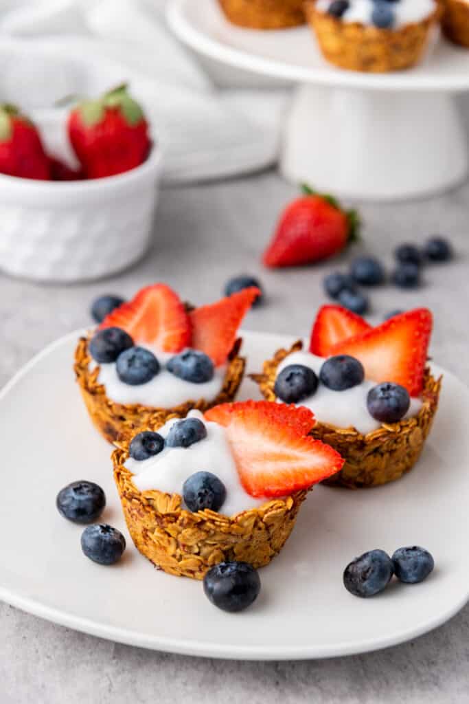Granola berry cups on a white serving plate, white ramekin with fresh berries and cake stand with granola berry cups in the background.