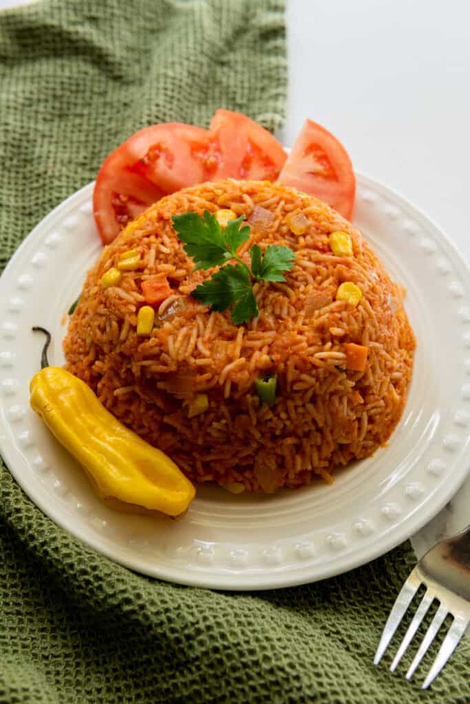 Jollof rice served on a white plate with fresh tomato wedges and habanero pepper, metal fork and olive green kitchen cloth on a white marble countertop.