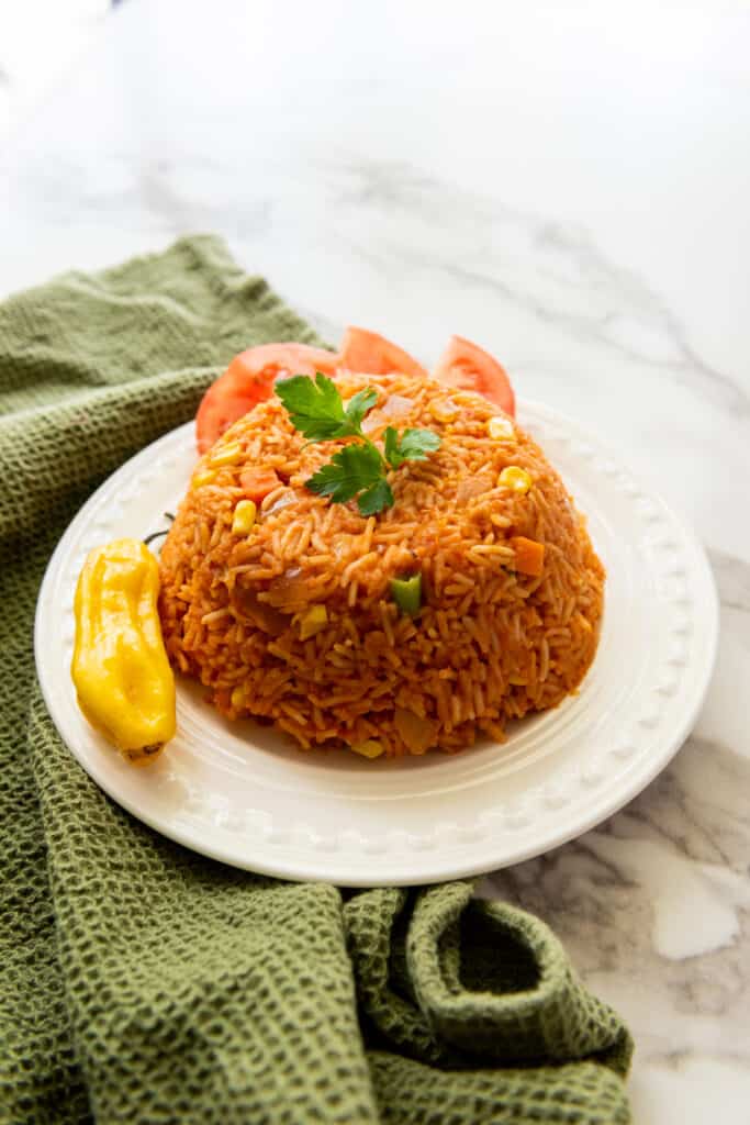 Jollof rice served on a white plate with fresh tomato wedges and habanero pepper, olive green kitchen cloth on a white marble countertop.