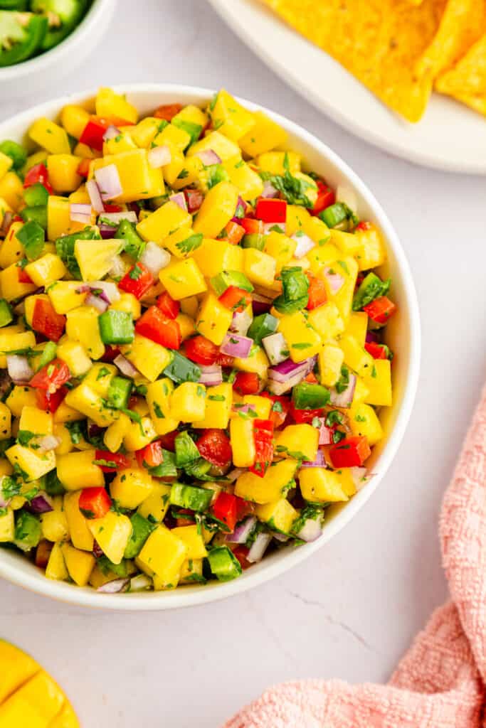 Close up of mango salsa with fresh mango salsa ingredients on a white kitchen countertop with a kitchen cloth.