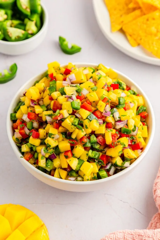 White serving bowl with mango salsa, fresh mango salsa ingredients on a white kitchen countertop with a kitchen cloth.