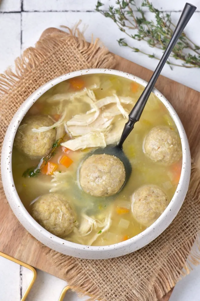 Overhead view of a bowl of Matzo ball soup with a metal spoon on a kitchen countertop.